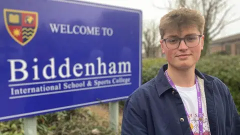 Alex Pope/BBC Finn, standing outside by a sign for Biddenham school, that is blue. He has short curly hair, is wearing glasses, and has a lanyard round his neck, a white patterned T-shirt and blue shirt on. There are trees and bushes behind him.