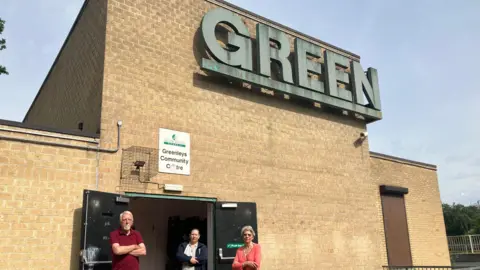 Amy Holmes/BBC A brick community centre with a large “GREEN” sign above the entrance, where three people stand in front of open doors behind a metal railing on a sunny day.