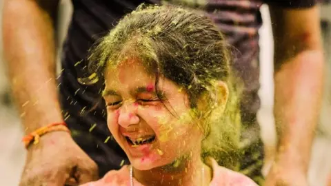 A young girl has yellow powder paint thrown at her during Holi festivities.