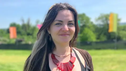 Addie P, a young woman, wearing a red chunky necklace with a brown cardigan. She has long dark brown hair with a blonde streak. She is smiling at the camera.
