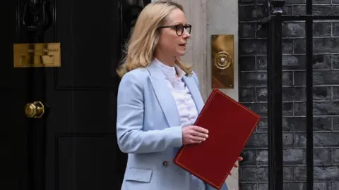 Lucy Rigby leaving 10 Downing Street, holding a red folder. She has long blonde hair, is wearing black glasses, and is wearing a light blue suit with a pale coloured shirt. 
