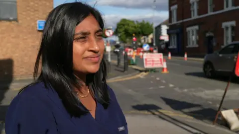 BBC Woman with dark shoulder length hair wears a dark blue top as she stands on a street with roadworks and a water leak in the background.