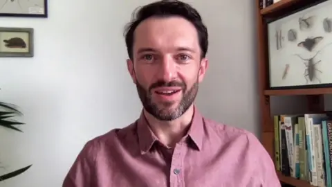 Dr Cockerill is sat in an office or study, talking to the camera. He has short, dark hair and beard and is wearing a dark red shirt. He is sat next to a book case with books and a picture of insects.