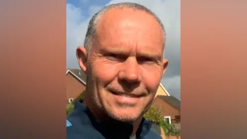 Steve Daniel A man with short shaved grey hair smiles at the camera. He is standing outside on a cloudy day. He is wearing a blue sports jumper.