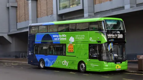 A double decker bus with a lime green front and blue back is pulled over on a bus stop. On the front of the bus, a display reads: 'Redbridge, via city centre'. It is the 300 service. On the side of the bus are the words "taking the NOx out of Oxford", "better air quality all over our city" and "electricity". In "electricity", the word 'city' is in bold typeface. 