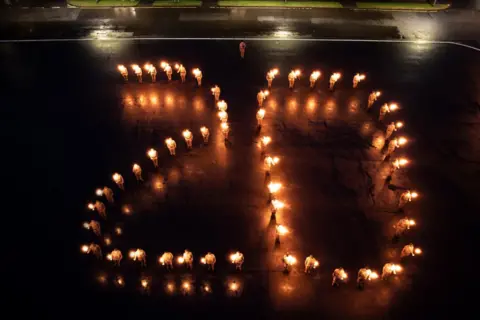 PA Media “Large group of uniformed individuals standing outdoors at night, each holding a torch, arranged in formation to create a glowing number ‘20’ on a dark wet surface, with a stone building illuminated in the background.”
