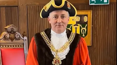 An AI generated image of Mayor of Gloucester Ashley Bowkett in red, white and brown ceremonial robes. It also features a hat and large gold chain. There is a red chair behind him.
