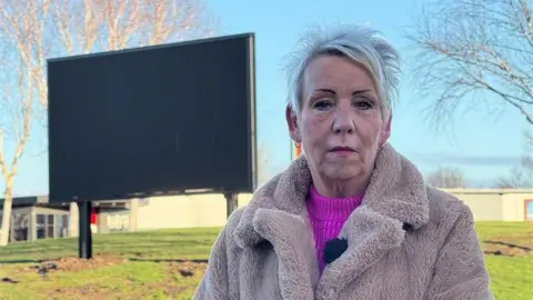 Lynn Appleby standing in front of the sign on the roundabout. She has spiky grey hair and is wearing a beige faux fur jacket.