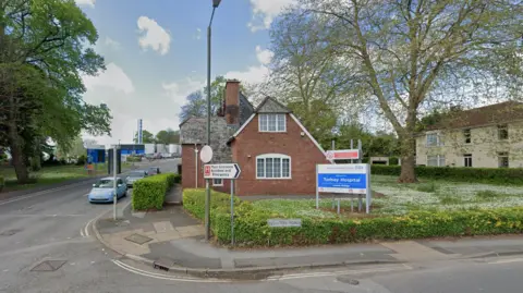 Torbay Hospital entrance pictured. The photo shows a blue welcome sign with "Torbay Hospital" written on it in white lettering. There is a small brick structure at the entrance and a road leading towards the main hospital building.