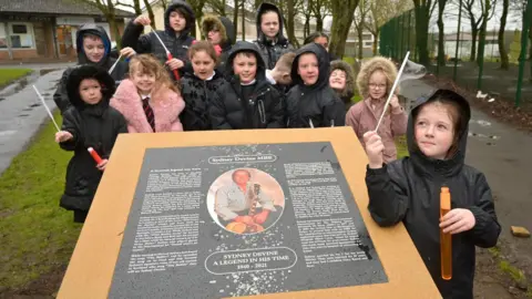 Children in waterproof clothing gather round a black memorial plaque to Sydney Devine MBE. The have tubes of soapy liquid with wands blowing bubbles in the wind