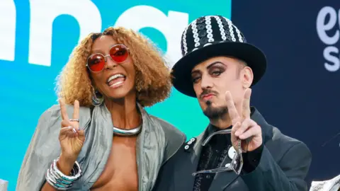 SENHIT and Boy George both make the peace sign. She wears a silver top, he wears a black suit and black and white hat