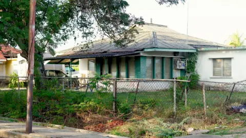 A white single-storey house with a grey roof and light green trim around the windows. The front lawn is surrounded by a low, wire fence.