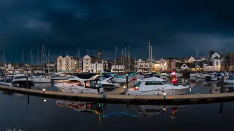 MANX SCENES Peel Marina at night, there are modern boats in the foreground and buildings in the background.