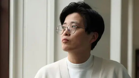 Yang Shuang-zi has short dark hair, wears thin, black rimmed glasses and wears a cream cardigan over a white t-shirt. She is staring out a window.