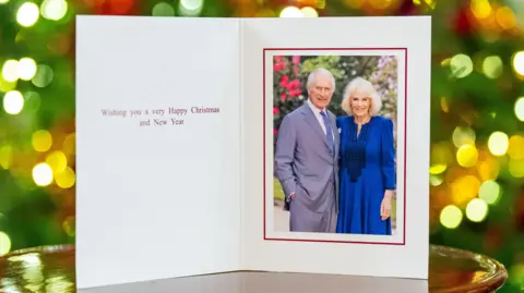 PA Media King and Queen standing side by side in photograph on card. The King - standing on the left - is wearing a grey suit and blue tie with his right hand tucked into his pocket, while the Queen wears a blue wool crepe dress. Card message reads: "Wishing you a very happy Christmas and New Year"