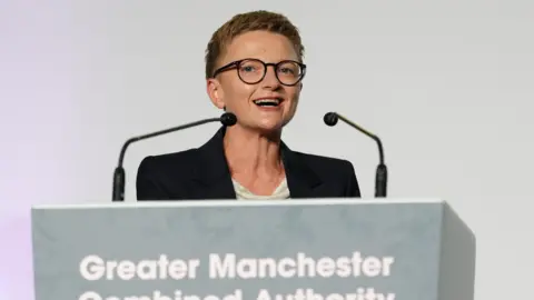 PA Bev Craig wearing a white top and black jacket stands on a platform speaking in front of a microphone.