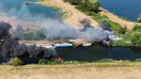 Drone Photos Sandy A photo taken from above by a drone shows thick black smoke billowing from a small burning boat in a dark river. Light smoke is coming from the charred remains of a small wooden structure on the other side of the water.