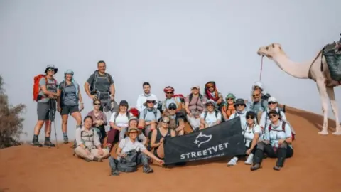 Vil Hoferica A group of men and women, some seated on the floor, some standing in the desert holding a black sign which reads StreetVet and there is a camel on the right-hand side