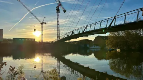 The footbridge is reflected in the water, the sun is low in the sky, and an aircraft has left a trail in the sky.