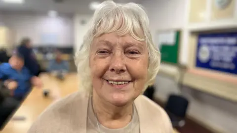 Brenda Maynard smiling for the camera. There are people behind her sitting at tables at the club.