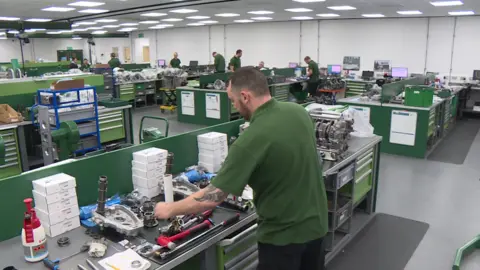 BBC A picture of work ongoing at Xtrac, with people engineering motor parts. A man stands close to the camera in a green T-shirt and is looking at a motor part.