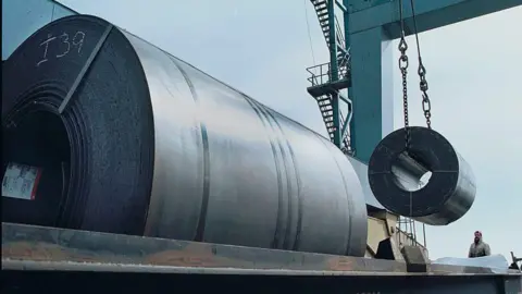 Workers use a crane to load a 60,000 pound coil of steel from Holland onto a railroad car for shipment to Ford Motor Company in Ohio from a warehouse at the South Jersey Port Corporation, March 6, 2002 in Camden, NJ along the Delaware River.