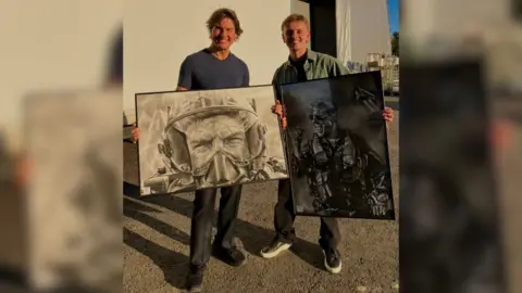 Wilf Elliott Actor Tom Cruise is standing with tattoo artist Wilf Elliott. They are each holding a large, framed black and white drawing of Tom Cruise. The actor is wearing a grey T-shirt and grey trousers, and the artist is wearing a green shirt and grey trousers. They are outside, standing on gravel with a large cream wall behind them.