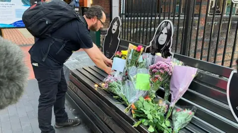 A man with dark hair touches tributes on the bench.