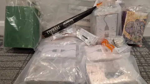 A number of items, including suspected drugs, seized by police, are sat on a table and on display. They are in clear plastic bags. behind them is a baseball bat
