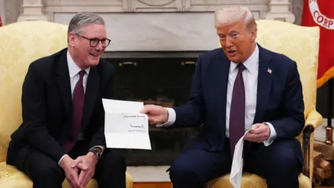 Two men sit beside each other on yellow chairs in the Oval Office. Donald Trump, with greying ginger hair and wearing a navy suit and purple tie, is holding a piece of paper in his right hand. Keir Starmer, with grey hair and glasses is on the left. He is wearing a dark suit and purple tie. 