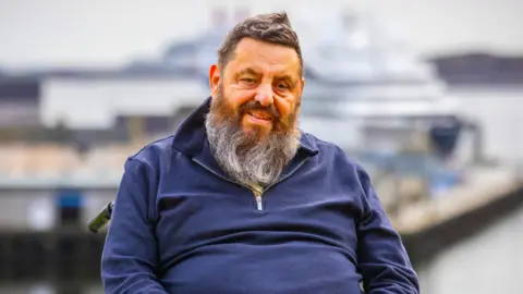 Suministrado Un hombre con una larga barba gris y pelo puntiagudo que lleva un jersey azul marino.