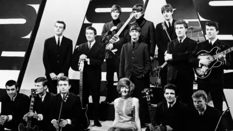 Getty Images A black and white photo of The Beatles and The Searchers, who pose at the top of a drum stage with Cilla Black and the band Billy J Kramer & The Dakotas sat on the bottom step. They are in a TV studio.