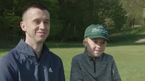 Wesley with his seven-year-old son Freddie on the golf course with trees and a bunker in the background. The father and son are wearing golf clothes. The pair are smiling. 