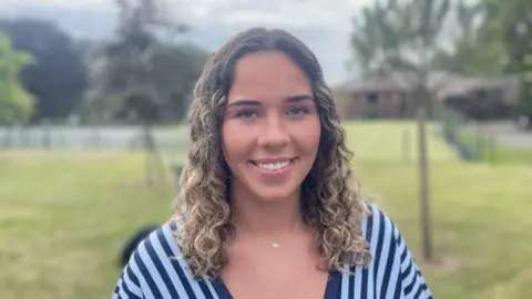 A woman with curly brown/blonde hair stairs at the camera. She wears a blue and navy striped top and a thin gold necklace. Behind her is an out-of-focus garden.