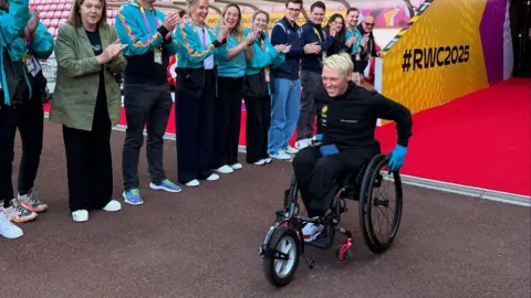 ChildFund Rugby A female wheelchair user with short, bleach blonde hair pulls into a sports stadium, with a line of people clapping her to one side.