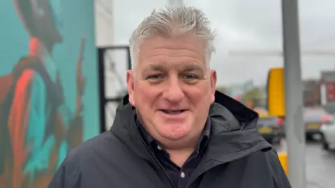 David is smiling at the camera. He is standing beside a set of traffic lights. He is wearing a black coat and has grey hair.