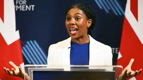Kemi Badenoch is wearing a white jacket and a blue top. She is speaking in front of a blue background.