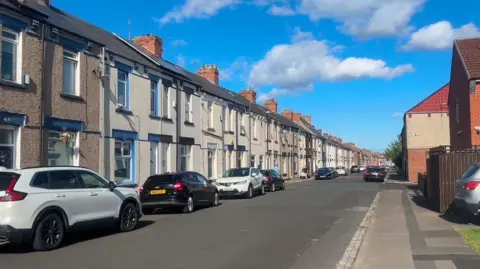 View of one side of a residential street of terraced houses with cars parked along the sides.