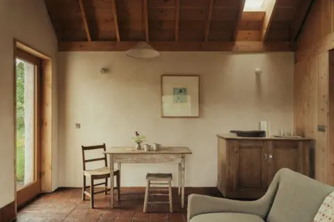Murray Orr The interior of a cottage, with a wooden table and chairs, with mugs and flowers on the table, a sideboard, sofa, painting on a wall, and window at the side.