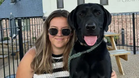 Katherine Selby Sharland and a black Labrador dog. She has long brown hair and sunglasses on and appears to be sat in a beer garden setting with wooden benches behind her. 