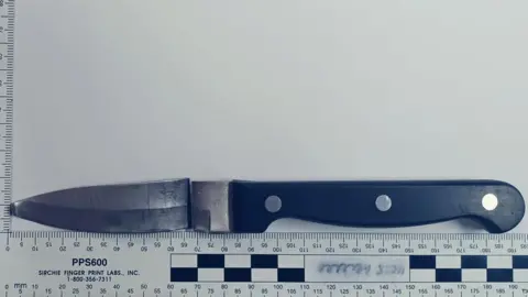 Crown Prosecution Service A black handled kitchen knife, the blade has been snapped away from the hilt but is placed above oit. Below is a ruler measuring the blade and handle to be 195cm long