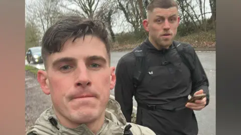 A man with short dark hair looks into the camera with pursed lips. Her wears a cream-coloured jacket. Beside and behind him is another man with close-cropped brown hair, the start of a beard and who wears a black jacket while he holds a phone in his left hand. The pair are on pavement to the side of a road, which can be seen behind them