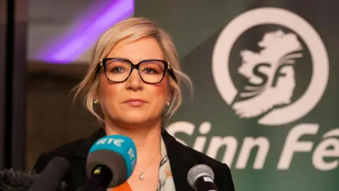 Michelle O'Neill with blonde hair tired up, wearing glasses. She is standing in front of a microphone.