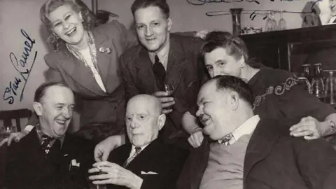A black and white signed photo of Laurel and Hardy's visit to the Plough Inn Barkston in 1947. Three men sit in the front row of the photo, with two women and a man behind then. A cabinet full of wine glasses can be seen in the background.