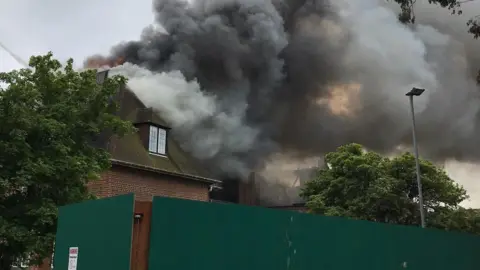 Tyne and Wear fire and Rescue Service Smoke billowing from Henderson Hall in Newcastle. Flames can be seen just above the roof of the building on its far side. The building is made of brown brick and has a dormer window in its roof. It is located behind green solid wooden panels.