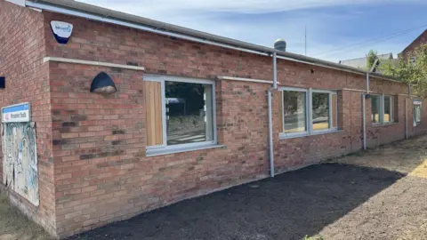 Shropshire Council A youth club building made of brown brick. The wall has been repaired and has a series of windows along its side wall with wooden shutter-type sections.
