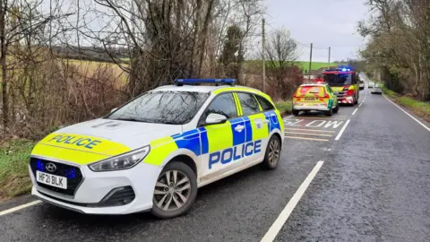 Dorset Police A police care is parked on a long country road. Further along the road, an ambulance and fire engine are also parked and their lights are flashing. Some other cars can be seen in the distance. Beside the road are trees and fields.