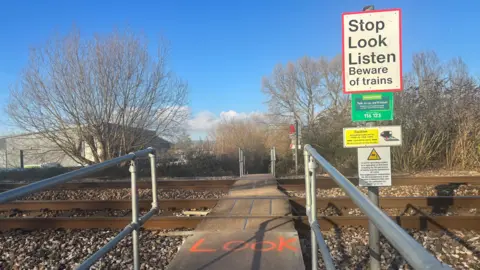 A foot crossing across a railway track. The word "Look" is spray painted on the floor and there is a sign to the right which says "Stop, Look, Listen". There is an industrial unit in the background to the left and trees to the right.