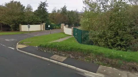 The road outside the headquarters of Heron and Brearley, with a dark green fence alongside two pale walls with a green gate between them.