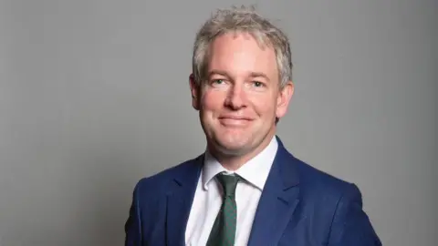 UK Parliament Danny Kruger wearing a white shirt, blue suit and green patterned tie. He has grey hair and blue eyes and is standing against a grey background, smiling at the camera.